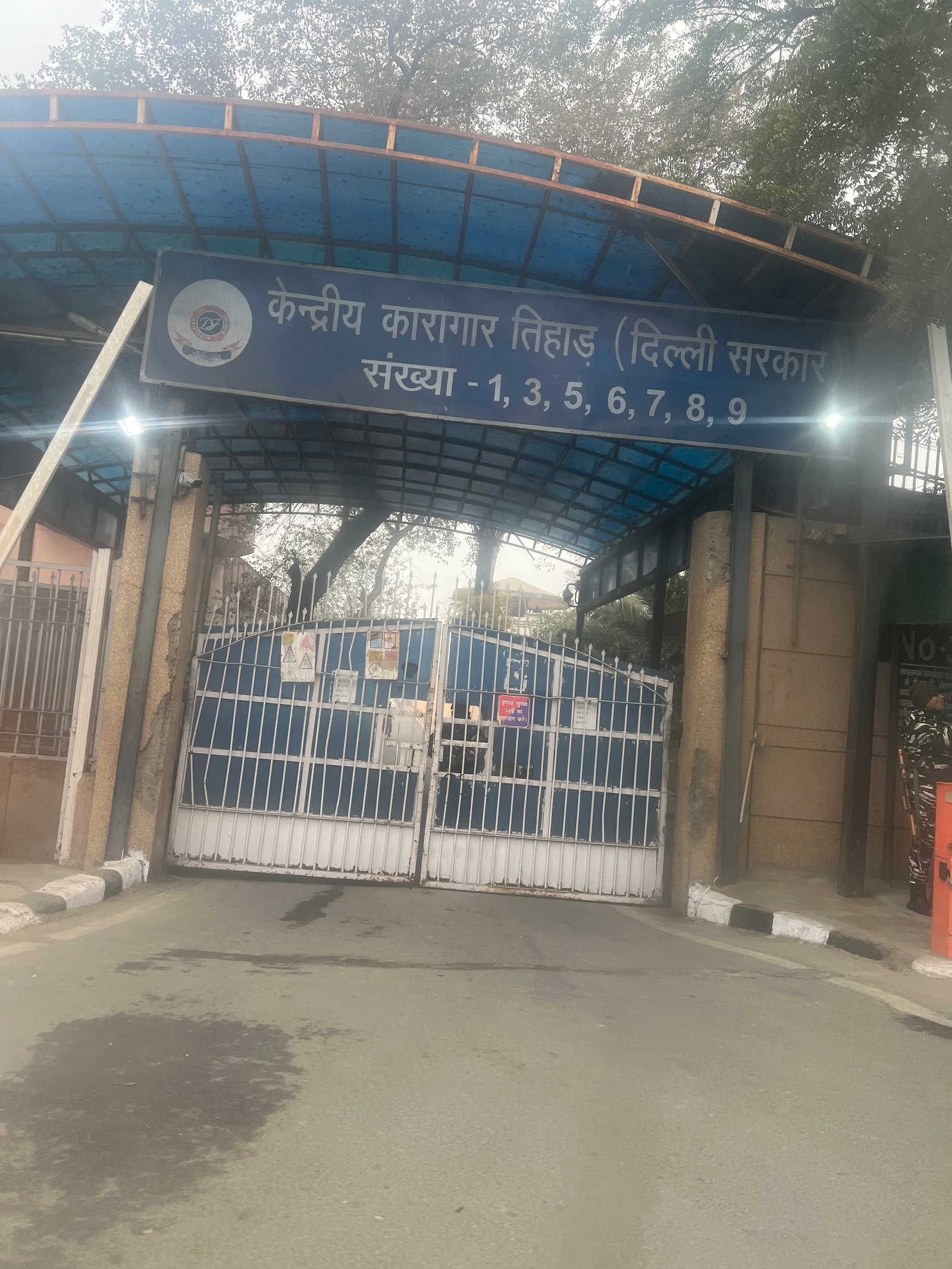 Modern photograph of a closed entrance gate at the Tihar Jail complex in Delhi beneath a blue Hindi sign.