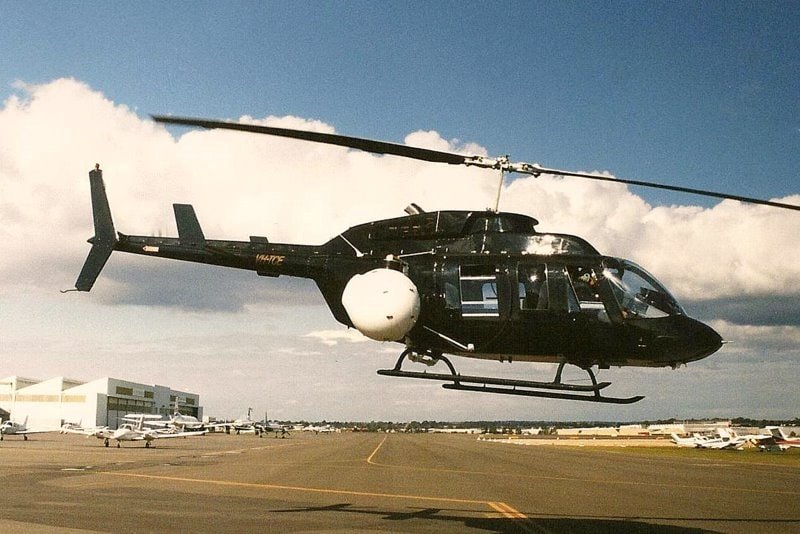 A black Bell 206L LongRanger helicopter flying low over an airport apron in 1998.